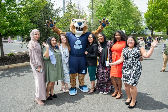 A group of students of different cultural backgrounds wearing colorful outfits and posing with the mascot Keanu with their hands raised