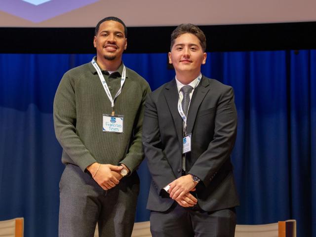 One young black man in a green pullover and a tan young man in a black suit stand on a stage in front of a projector screen