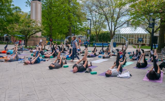 A yoga lesson taking place in front MSC patio. Students are laying on yoga mats in the same pose holding their right leg up.
