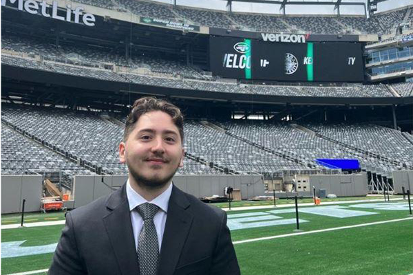 A young man with a beard, wearing a business suit, stands on the field at Metlife Stadium.