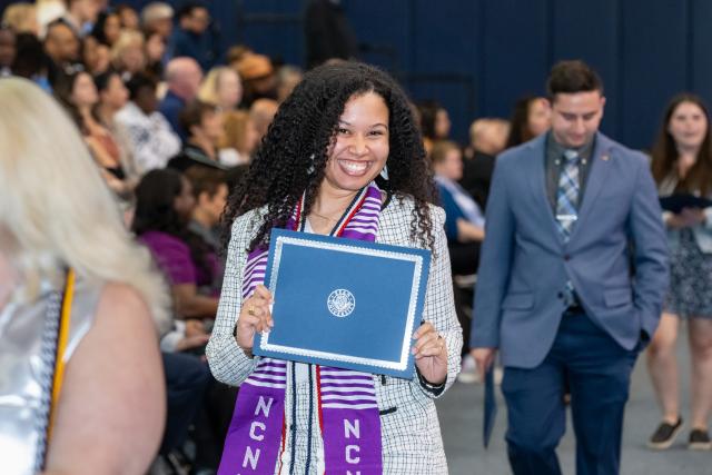 A woman with curly long hair, purple stole, holding a Kean certificate smiling at the camera