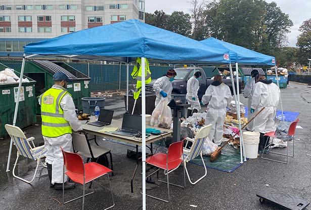 A group of people in protective gear, under two blue tents, sort trash on a tarp.