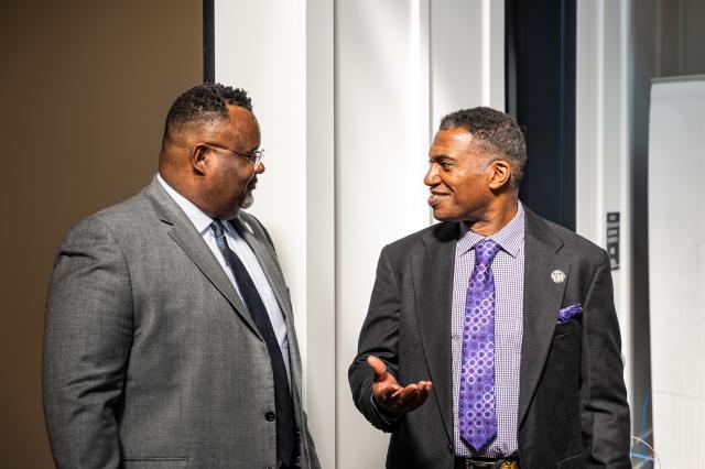 Two black men in suits, one sporting a gray suit, the other a black suit with a purple tie, speaking to each other.
