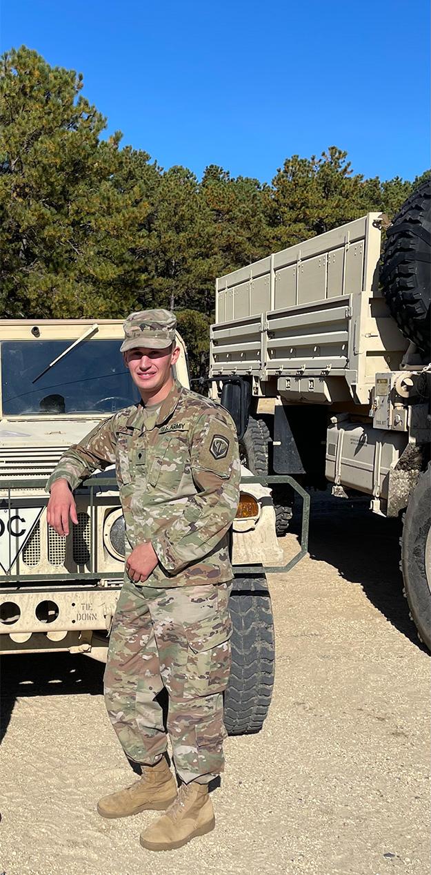 A young man in army fatigues leans against a white Jeep.