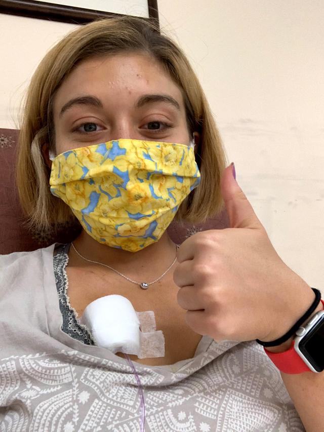 A young woman, with a chemo port in her chest and wearing a yellow face mask, gives a thumbs up