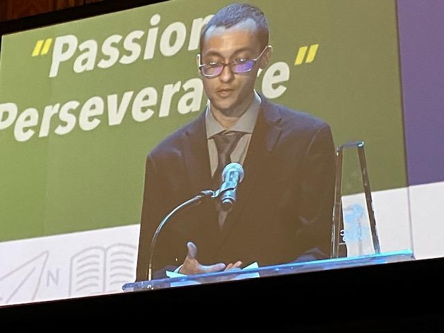 A close up shot of Xavier Amparo, a white male with buzzed brown hair, a peach fuzz beard and mustache, wearing black glasses, a black suit jacket, a brown button-up with a brown tie, standing in front of a podium, speaking into a microphone. Xavier is looking down, and reading from a piece of paper.