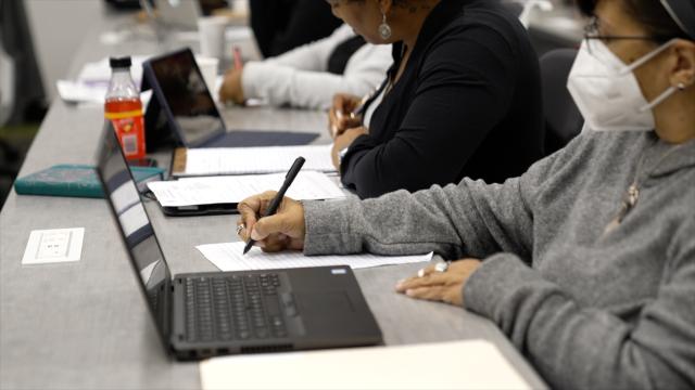 A woman, wearing a face mask, takes a note as she views a laptop; more people work beside her.