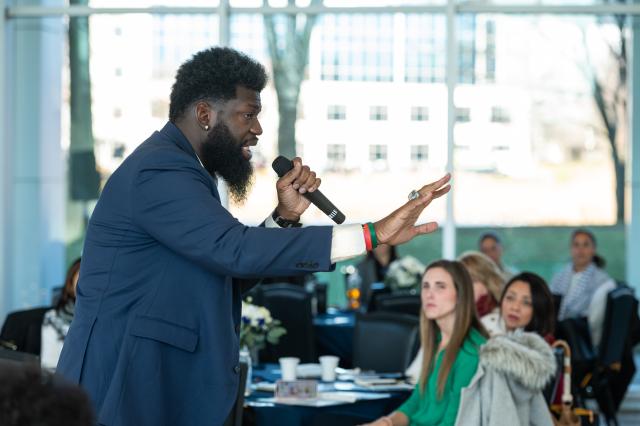 A Black man with a beard is seen in profile, speaking into a microphone and gesturing.