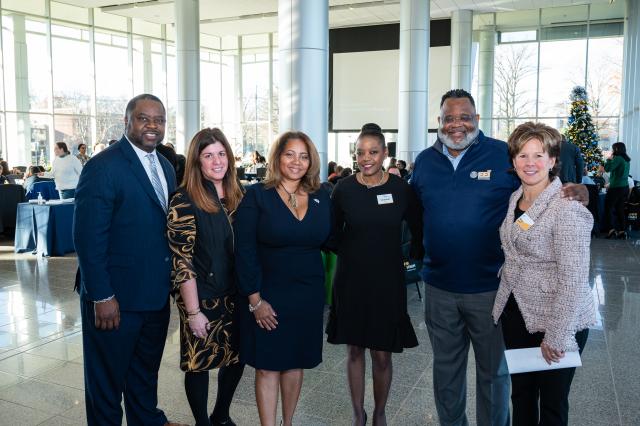 (L-R) A Black male with black hair and facial hair wearing a navy suit, a White female with long, brown hair wearing a long-sleeved brown and gold dress, a Black female with light brown hair, wearing a long-sleeved navy blue dress, a Black female with dark brown hair in a bun, wearing a long-sleeved black dress, President Lamont Repollet, a Black male with curly short black hair, wearing gray pants a navy blue top, and a white female with short brown/blonde hair, wearing a white blazer with black pants