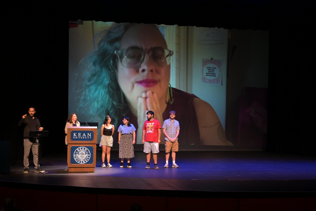 An interpreter, professor and four students stand on stage, with Leah Lakshmi Piepzna-Samarasinha on a large screen behind them