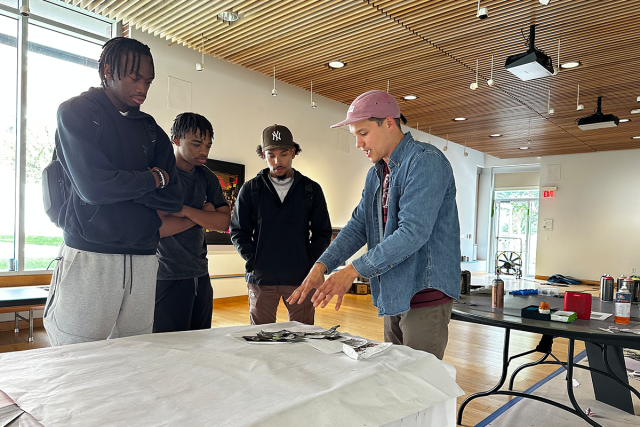 Kean alumnus and artist Ricardo Roig, with students, points at art materials on a table.