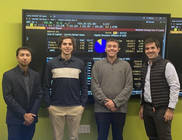 Two finance professors and two students post in front of a screen displaying financial markets information.