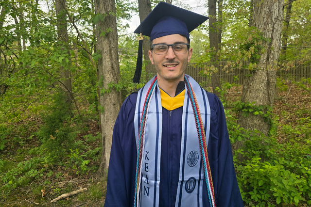 Kevin Hannon, in his blue Kean graduation cap and gown, stands in front woods.
