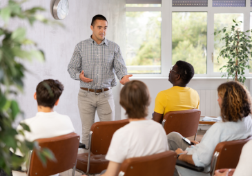 A man is standing and speaking to a group of students that are sitting in chairs in front of him. 