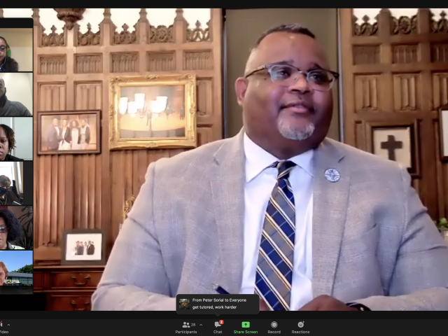 A screen shot of a Zoom event. Dr. Lamont Repollet speaks to First-Generation Students via Zoom. He is a Black male wearing a white shirt, gray suit jacket and striped tie, sitting in front of a wood-panel wall. Six people appear in Zoom boxes to the left of the frame.
