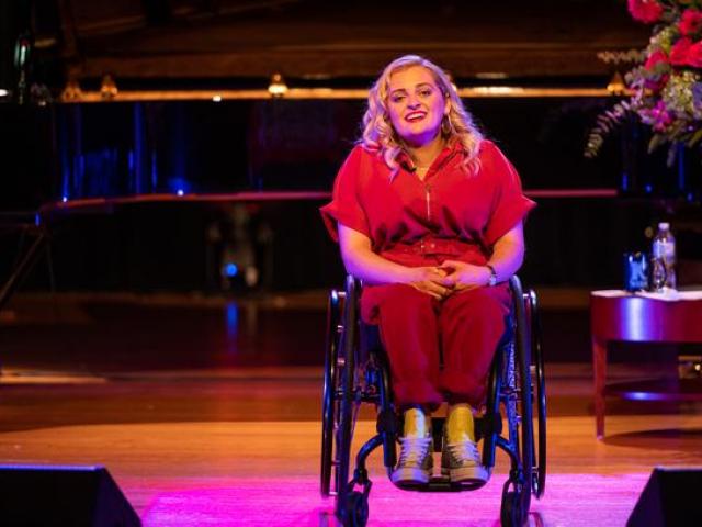 Ali Stroker, a female with curly blond hair, sitting in her wheelchair on stage. She is wearing a red jump suit. Head tilted, singing. Stage is lit pink. Piano and flowers can partially be seen behind her.