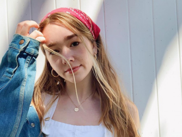 Holly Taylor in a red bandana, white shirt and jeans jacket, in front of a white wood frame wall.