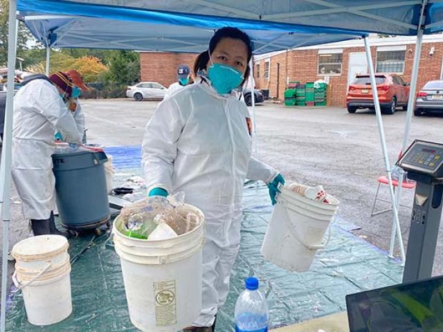 Kean professor Dongyan Mu in protective clothing hold two pails of sorted trash under a blue tent.