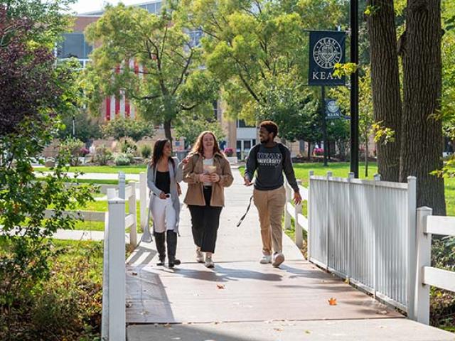 Three students, two women and a man, walk over a white bridge on Kean University's Union campus