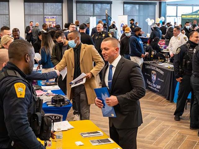 Attendees at a crowded recruitment event talk with law enforcement officers who are manning information tables.