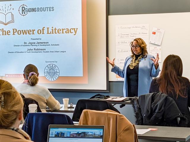 A brown woman with short, blonde hair and glasses wearing a light blue colored coat presenting a PowerPoint to a classroom full of students on The Power of Literacy.