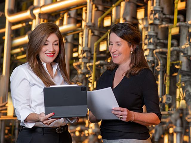 Two businesswomen standing before a wall of machinery and pipes; one holds a tablet, the other holds papers.