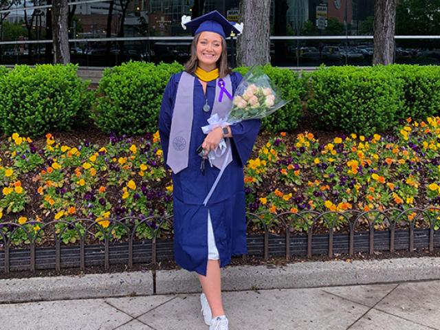 A young woman in blue graduation cap and gown, holds flowers as she stands in front of floral landscaping.