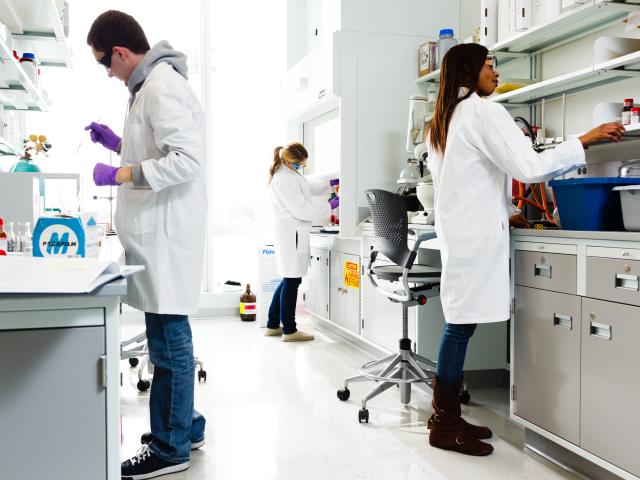 Three people with white lab coats and goggles work in lab.