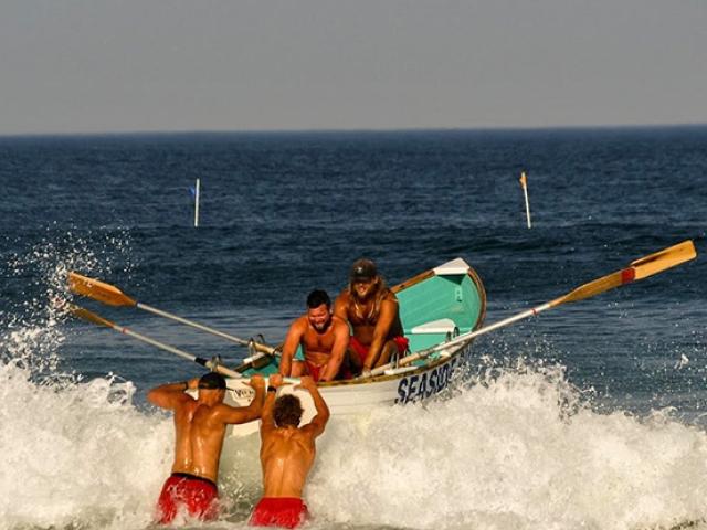 Four lifeguards, two in a rowboat and two pushing it, row out over ocean waves.