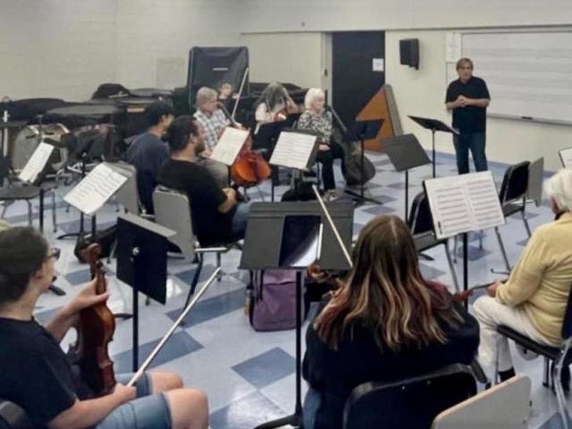 A conductor stands in the front of a rehearsal room filled with orchestral musicians.