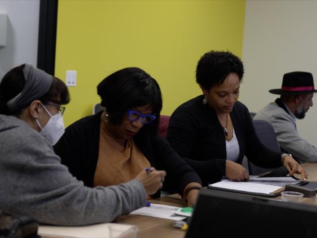 Three women and a man sit along a long desk, working together on computers.