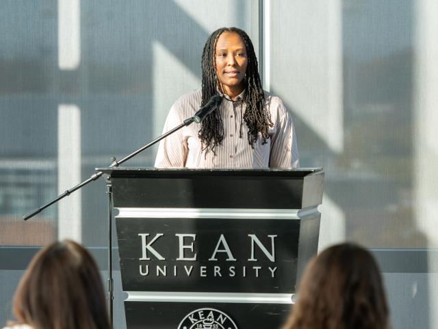 Chamique Holdsclaw, a Black female with long, black dreads standing behind a black podium that says Kean University on it in white. There is a black microphone in front of her as well. Chamique is wearing a cream-colored buttoned-up shirt, and is looking into the crowd.
