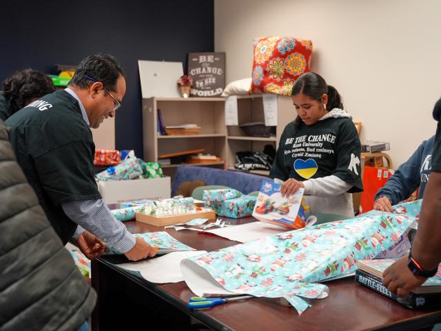 Three diverse people wrap Christmas gifts around a long table