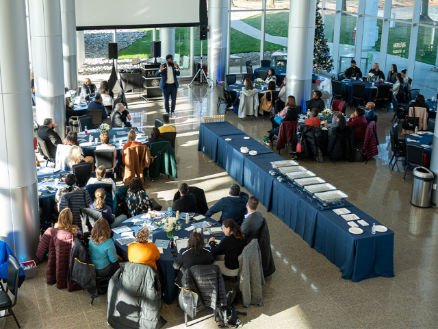 An overhead shot of a conference space with glass walls. A speaker is at the microphone, and round tables are filled with participants.