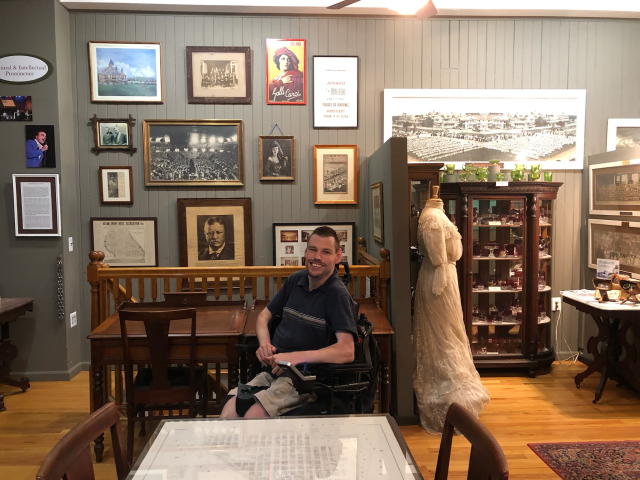 A white male wearing a dark t-shirt and tan shorts, in a wheelchair, pictured in front of old-time pictures and artifacts