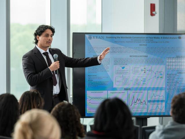 Eric Landaverde, in a jacket and tie, points to a his research on a large screen.