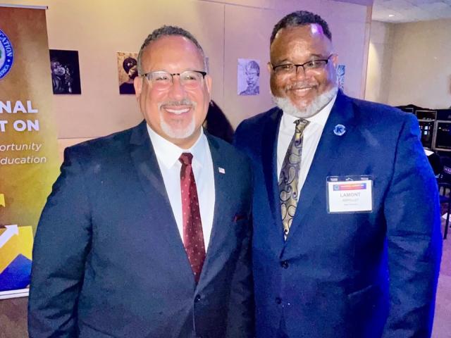 Two mean wearing blue suits; one Hispanic, one Black, stand shoulder to shoulder, smiling.