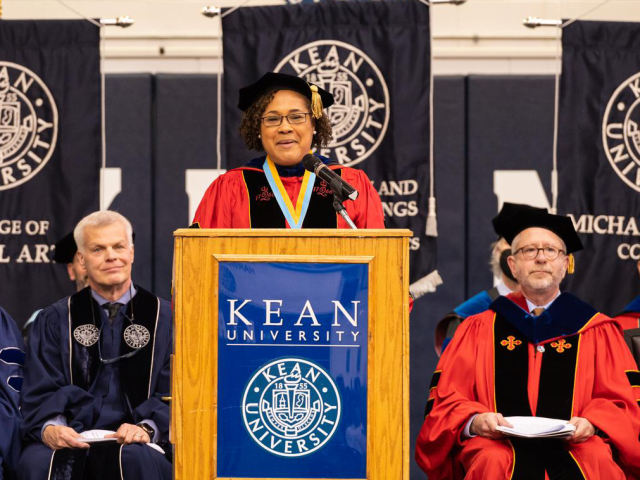 Rajade Berry-James, in a red commencement robe, speaks from a Kean podium at Harwood Arena.