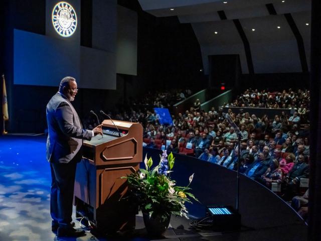 A side view of Kean University Repollet at the Wilkins Theatre podium, with the audience in front of him and the Kean seal projected on a wall.