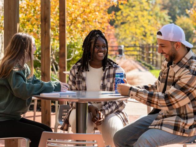 Three students at a round table laugh. A bridge and falll foliage are behind them.