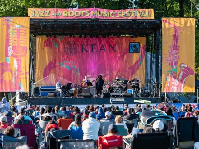 A woman sings on an outdoor, colorful stage, with an audience sitting on lawn chairs