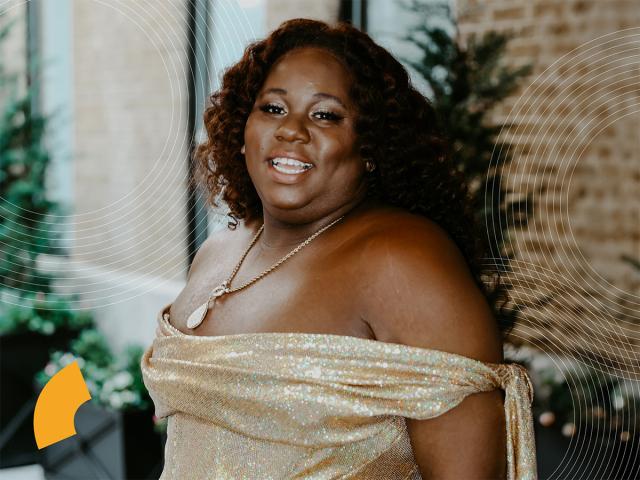 Alex with a golden dress, hair down and a big smile. They have a necklace and pine trees in the back
