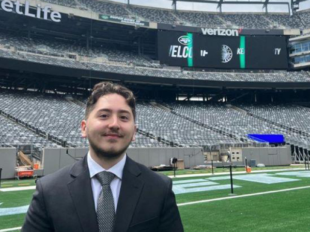 A young man with a beard, wearing a business suit, stands on the field at Metlife Stadium.