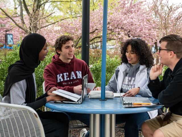 Four students, two women and two men, sit at a table with their laptops, outdoors on Kean's Union campus