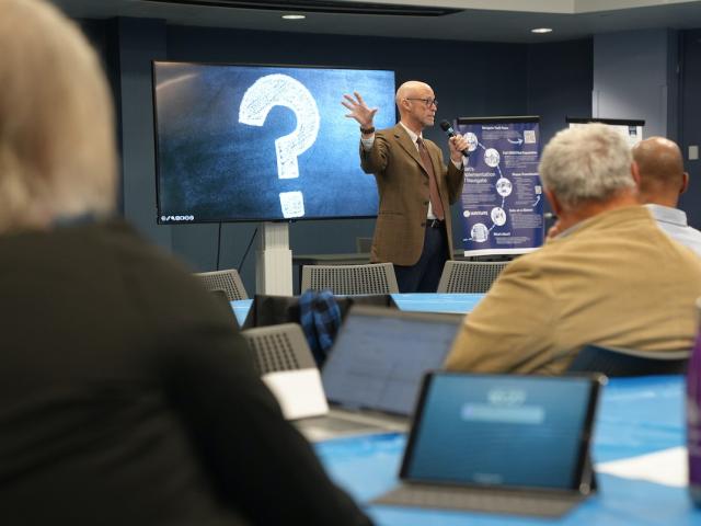 Dr. Henning at the front of a room gestures, while an audience seated at tables listens.