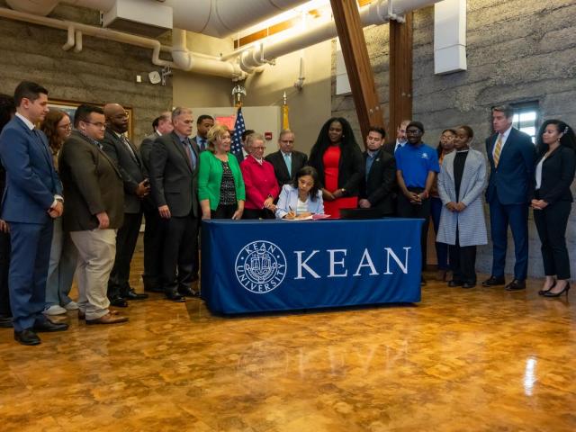 Acting Governor Tahesha Way at a table with the Kean seal signs a bill while a group of students, lawmakers and mental health advocates look on.