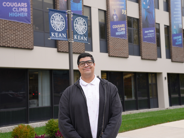 Gabriel Serrano, wearing a black jacket over a white shirt, stands in front of a Kean University building.