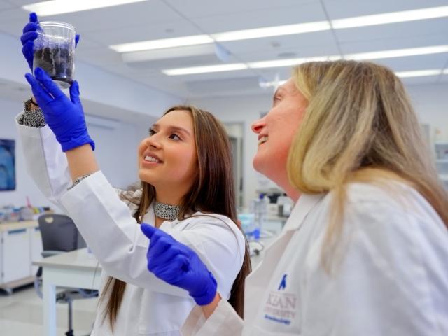 A female student holds a beaker aloft as a female professor looks on.