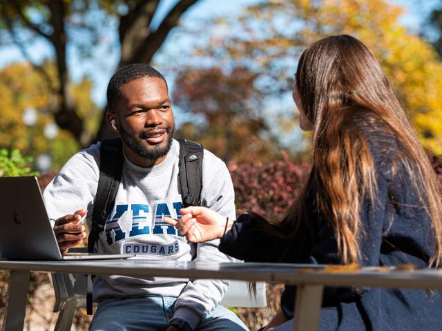 Student in a Kean sweatshirt seated at table outdoors and speaking with another student