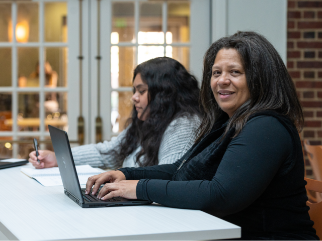 Two students seated next to each other, one working on a laptop while looking at the camera